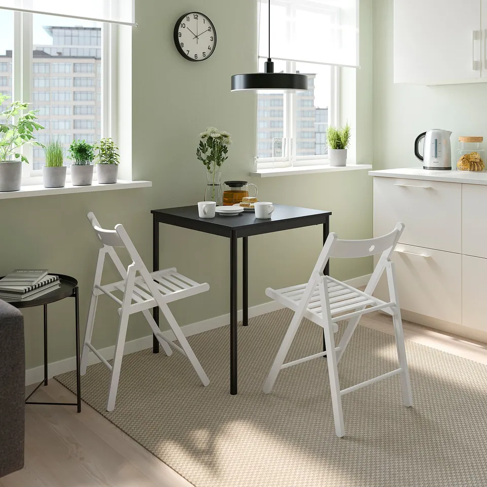 Small dining area with black table, white chairs, and green walls. Table set with cups and a vase. Chairs fold for space-saving.