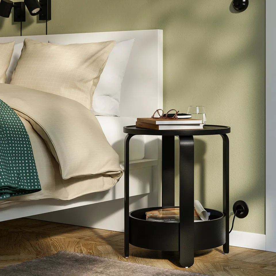 Black BORGEBY side table with shelf beside bed, holding books and glasses.