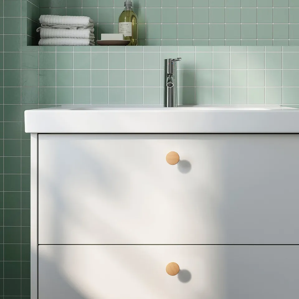 White vanity with wooden knobs, chrome tap, green tiles, stacked towels.