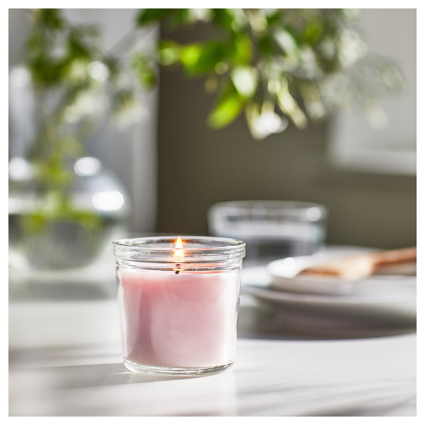 A romantic pink scented candle in a clear glass jar, lit, with green plants in the background.