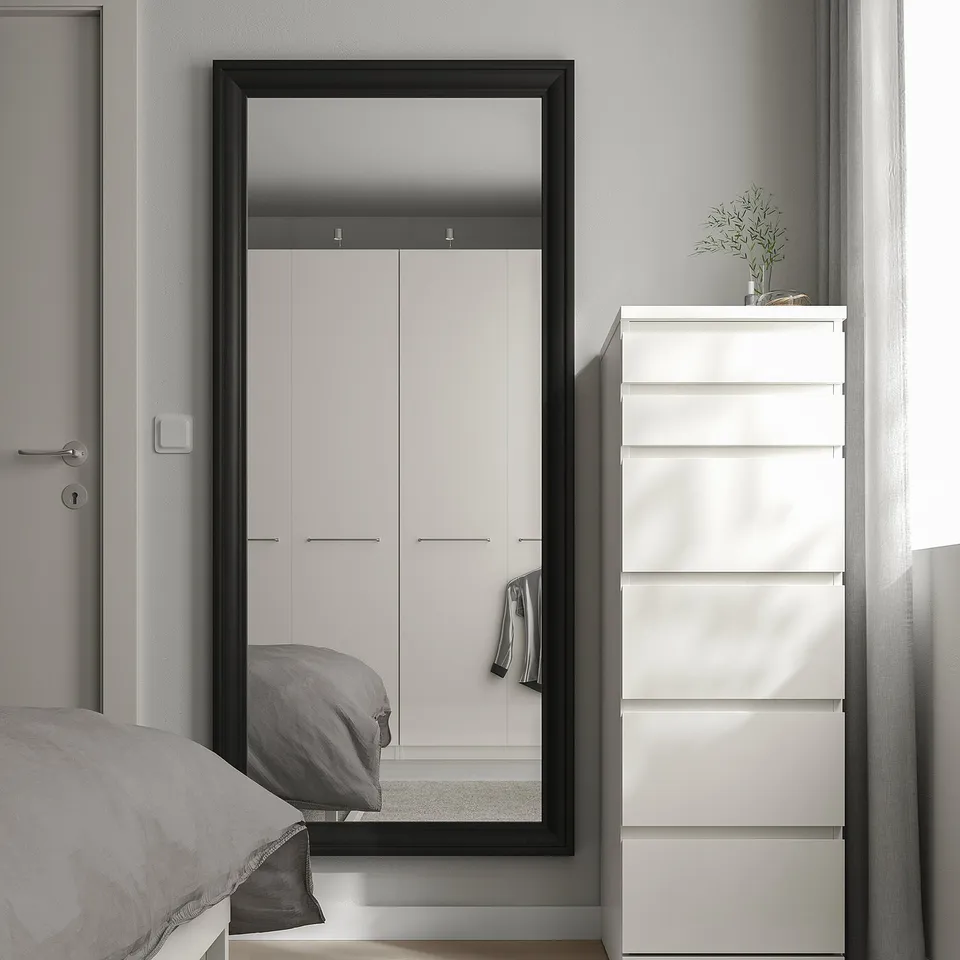 The TOFTBYN black-framed mirror stands tall between closet doors, reflecting a bed and dresser, adding a sleek, classical touch to the room.