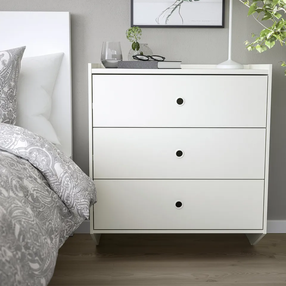 White RACKNÄS chest with drawers, wall-mounted beside bed. Top: glass, vase, glasses, book.