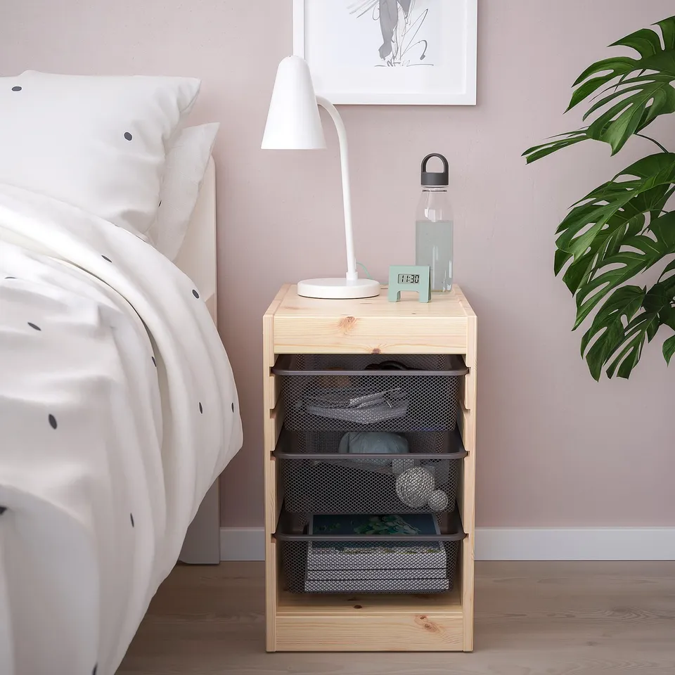 Small wooden TROFAST bedside table, light wood, with mesh storage. Holds a lamp, water bottle, and clock. Beside a white bed with a large houseplant.