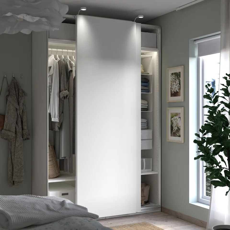 Bedroom with open white PAX wardrobe revealing clothes, shelves, and storage; grey walls and window with plant.