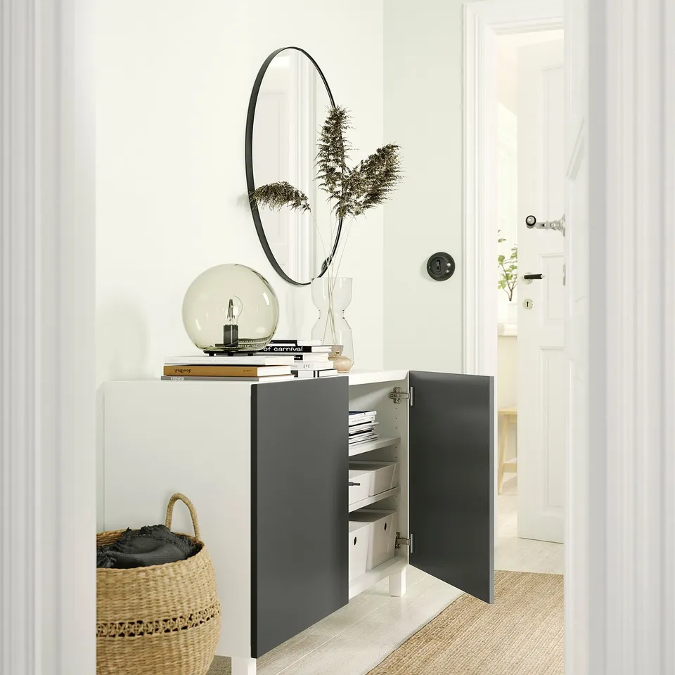 Modern white and grey BESTÅ storage unit with black-framed round mirror, decorative light bulb, and dried plants.