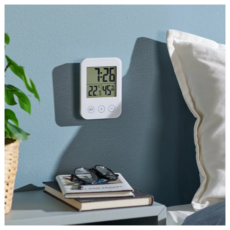 Bedside, white clock displays time, temp, and humidity on a blue wall near a plant and books.