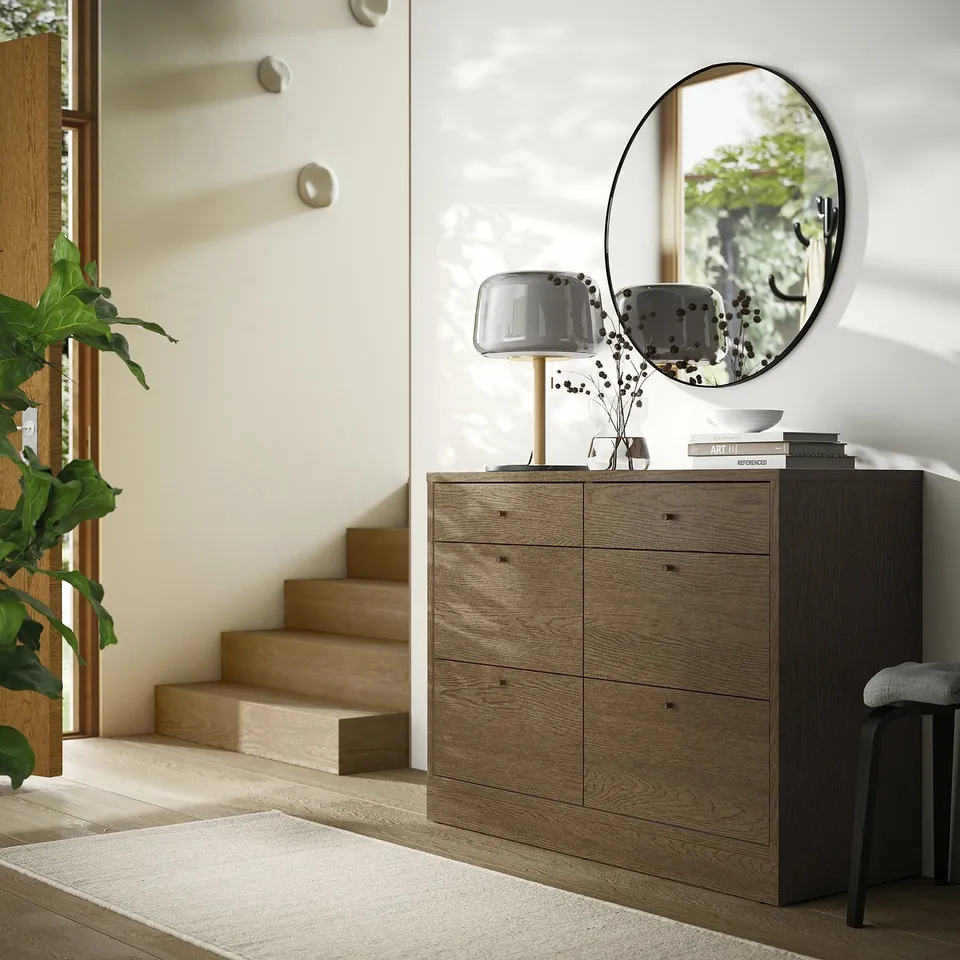 Modern entryway with TONSTAD chest, light wood, round mirror, grey lamp, stacked books, bowl, stairway, plant.