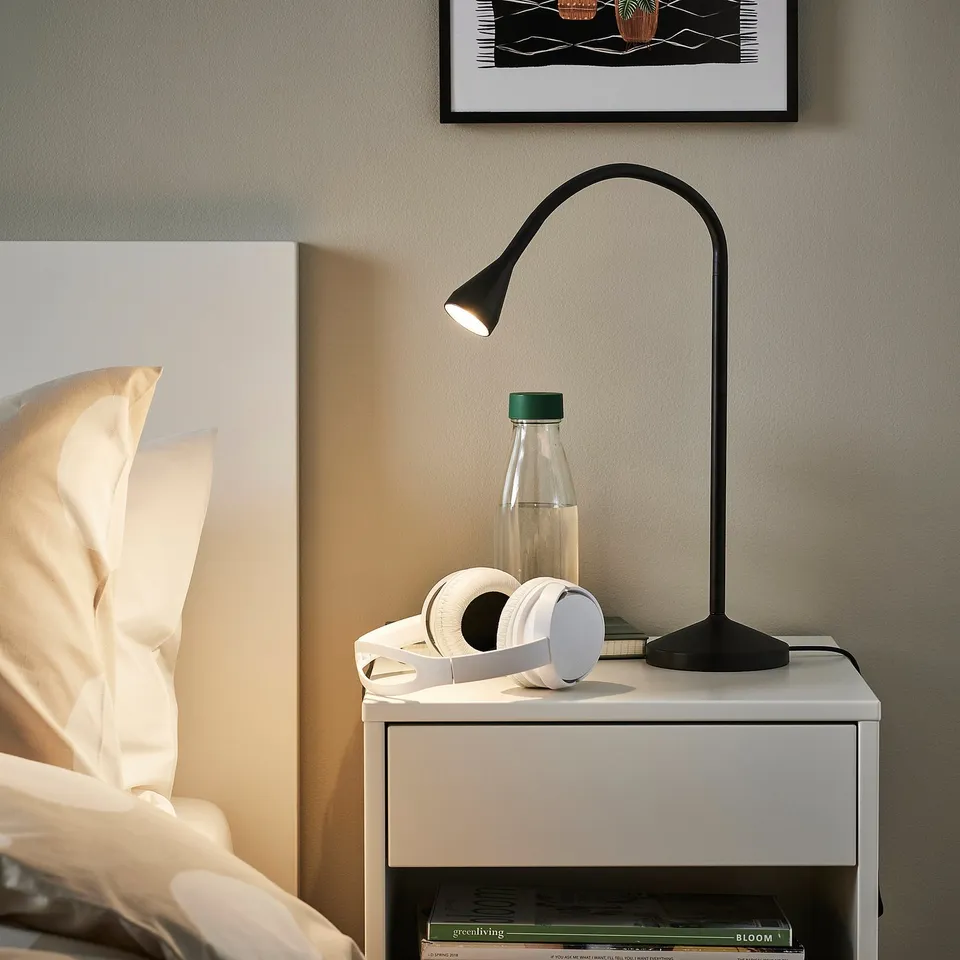 Modern bedroom with white bedding, NÄVLINGE lamp, headphones, water bottle, and books.