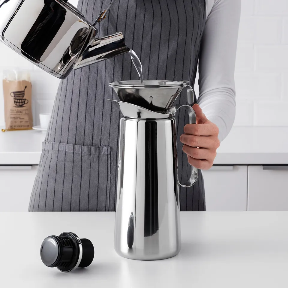 A person pours water into a metallic carafe on a white countertop.