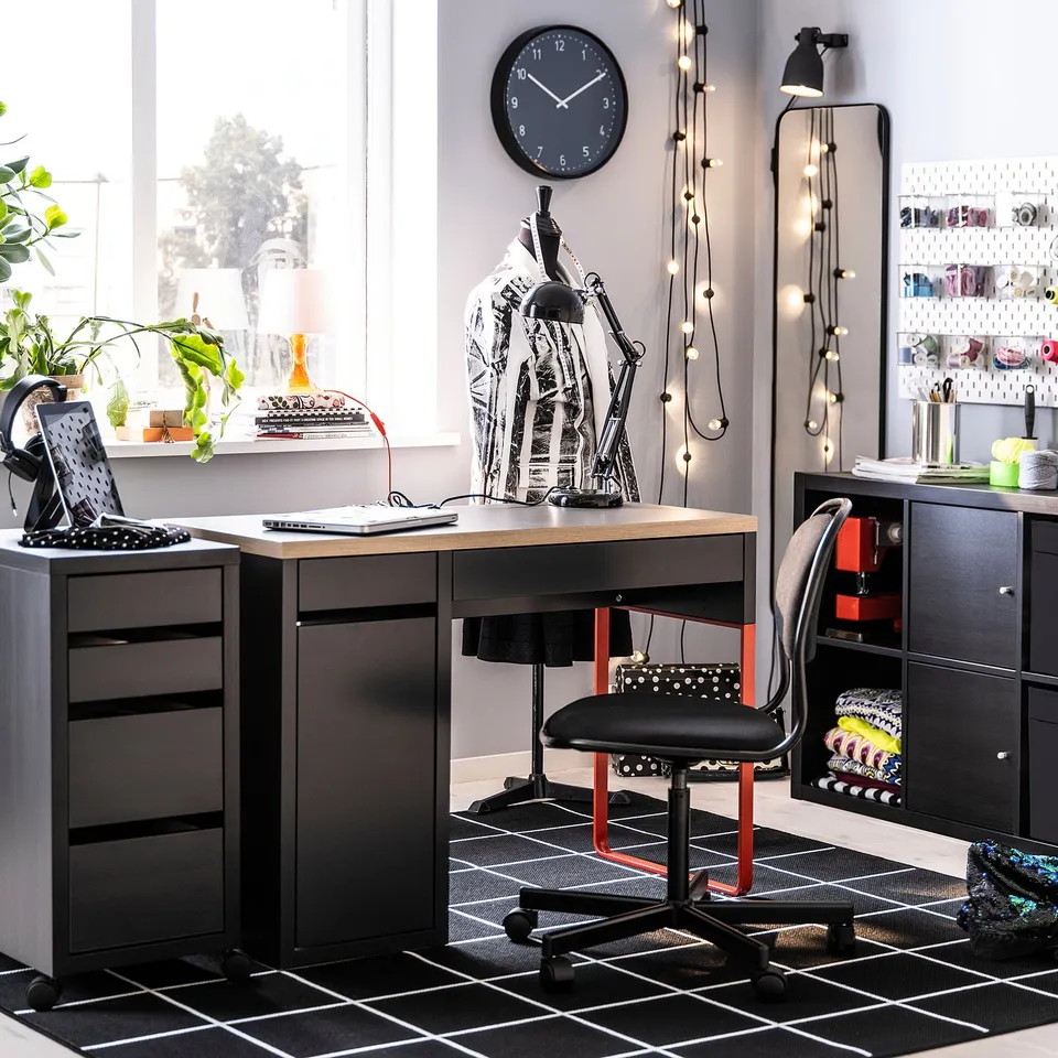 Well-organized home office with a black MICKE desk, red chair, and storage units. A large window, plants, and decorative lights for a cosy workspace.