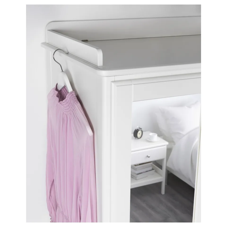 A white clothes rail holds a pink garment, mounted on a white wall next to a white bed and a bedside table with a clock.