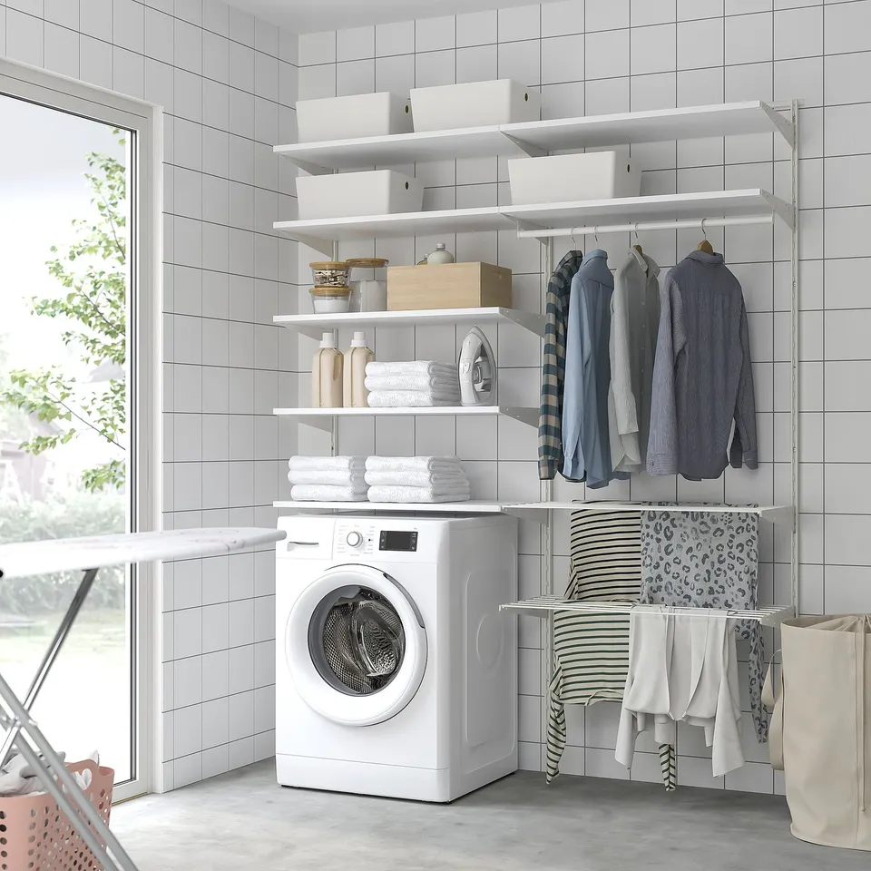 A tidy laundry room with white BOAXEL storage shelves holding folded towels, bottles, and clothes, a white washing machine, and hanging shirts.