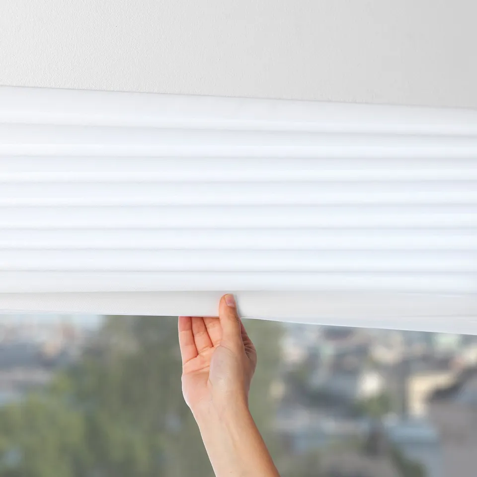 A hand pulls a white window blind downward, blocking light and offering privacy. The pleated fabric dims daylight, creating a cosy indoor atmosphere.