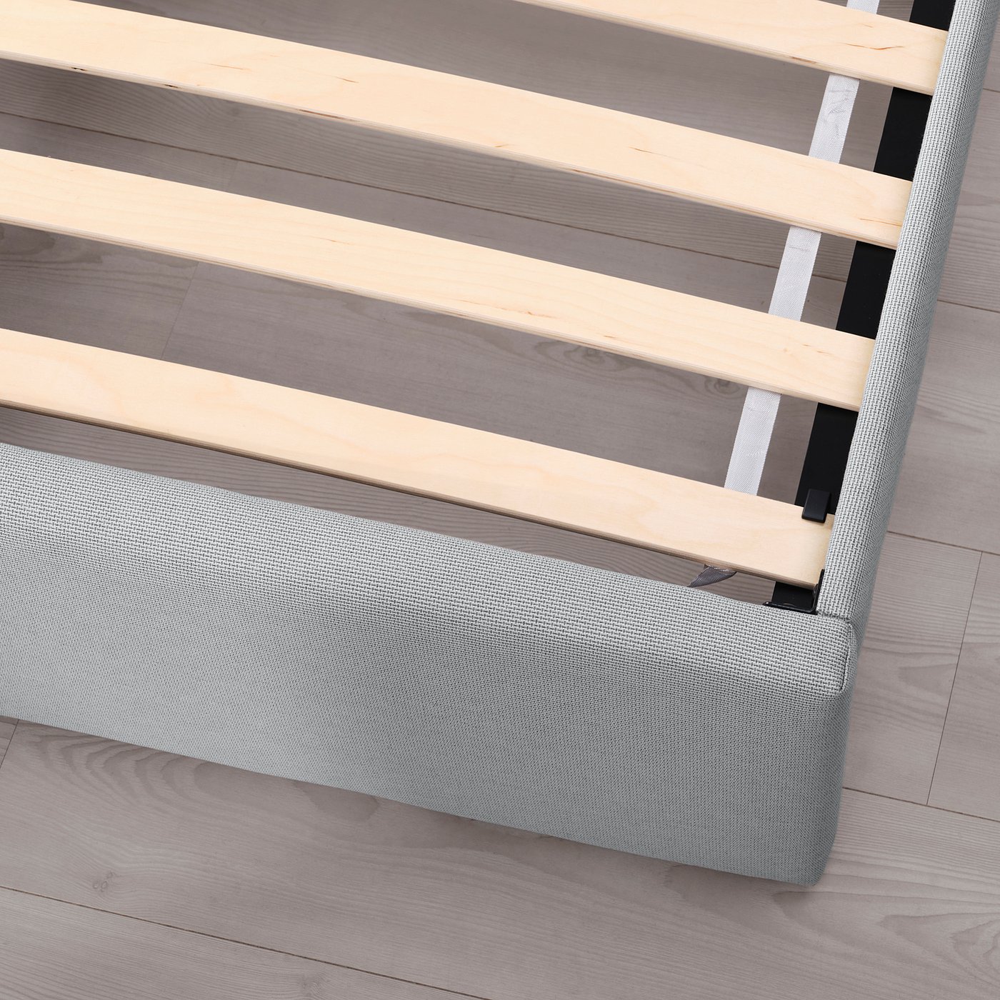 Closeup of upholstered bed frame with slatted wooden base and grey fabric. Storage boxes on castors are visible.