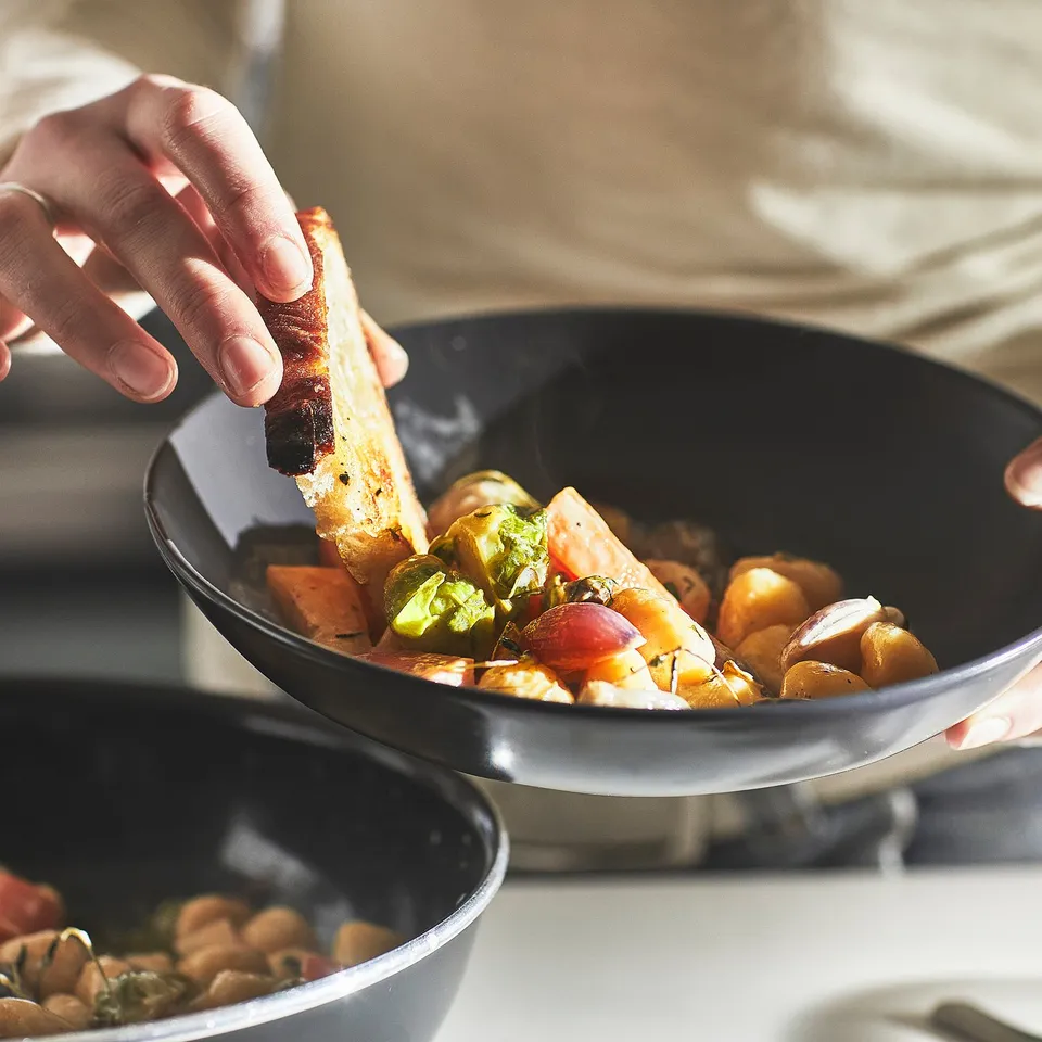 A hand holds bread over a frying pan with colourful vegetables, showcasing sautéing in a HEMLAGAD pan.