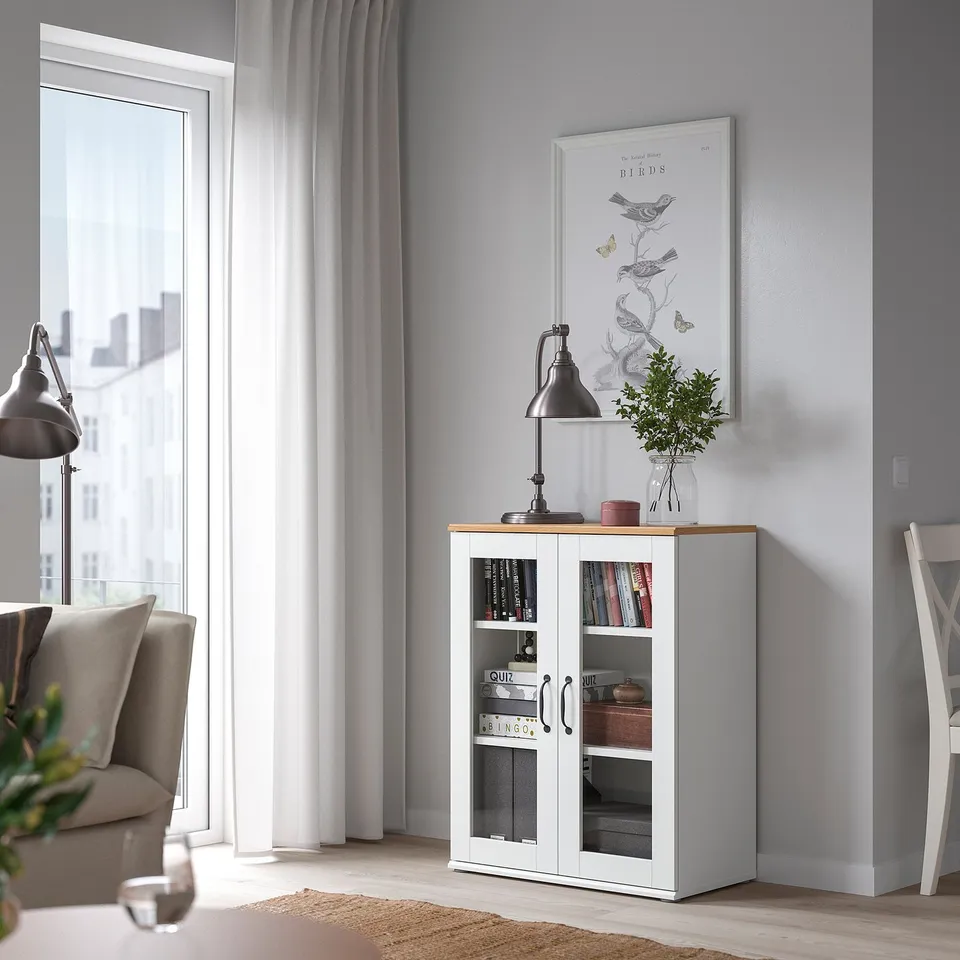 Modern room with glass cabinet, books, lamp, and plant by window. Grey walls, light floors, cosy.