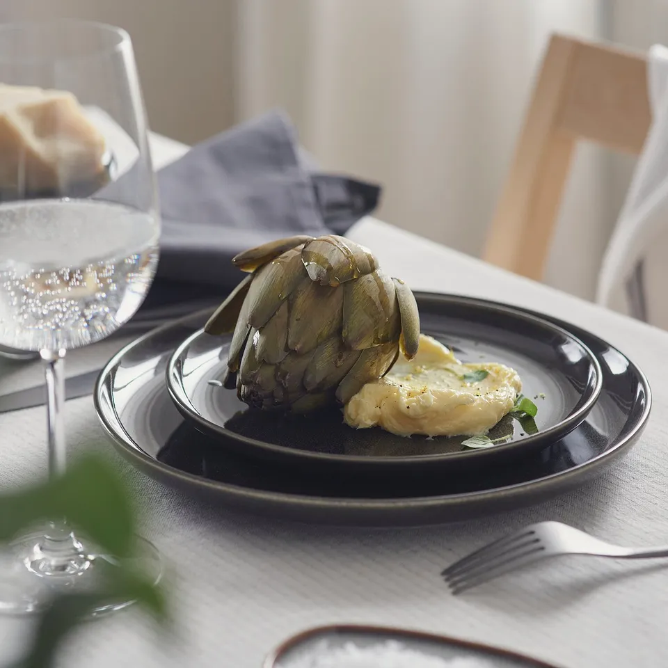 Dark-grey glazed side plate holding artichoke with sauce, on a table with a wine glass, tray, and cutlery, showcasing its crafted look.