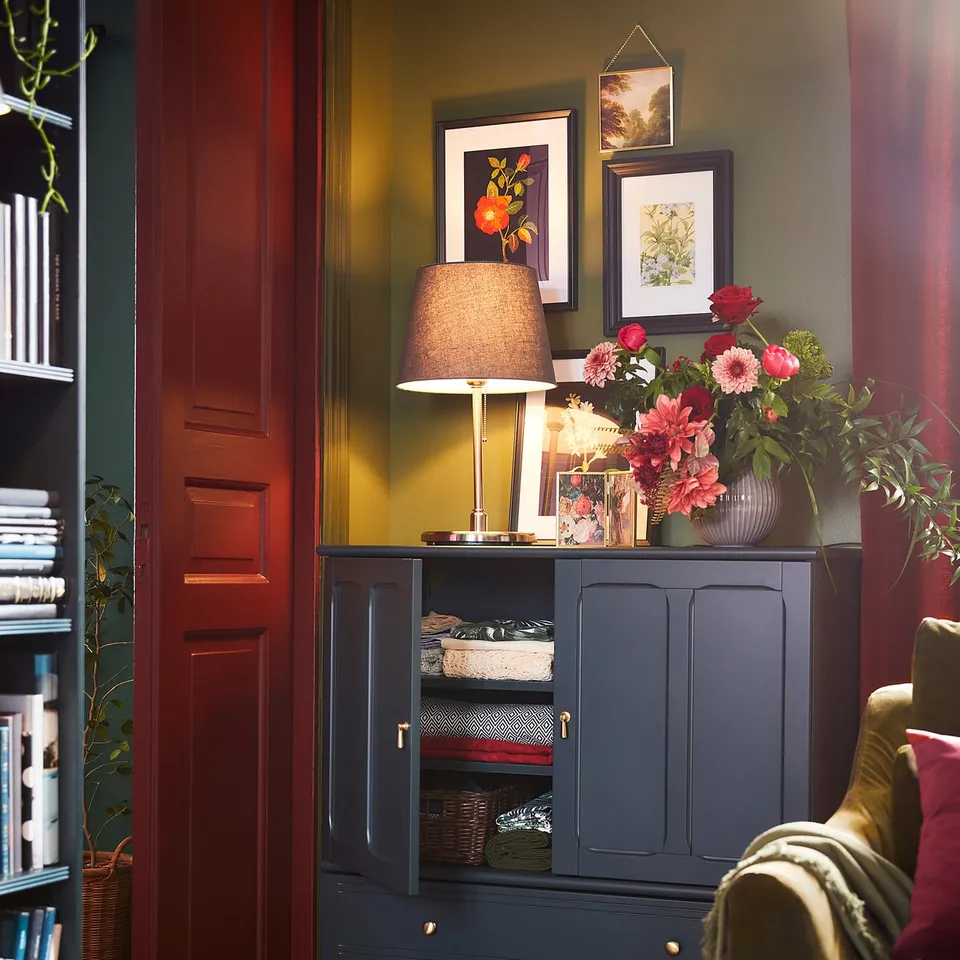 A cosy room with dark blue SKOTTORP lamp, green walls, wooden floor, bookshelf, and armchair with red pillow.