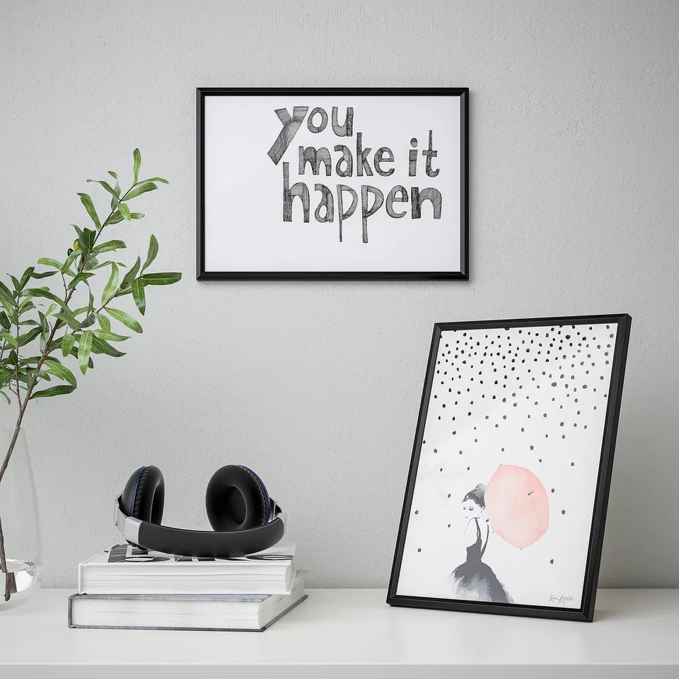 Minimalist desk setup with motivational art. Frame displays you make it happen poster, standing on books near headphones.