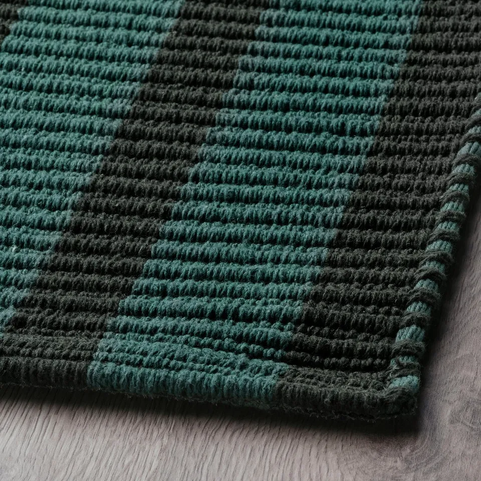 Dark green and black textured rug, laid on wooden floor. Visible stripes and dense weave suggest high quality and durability.