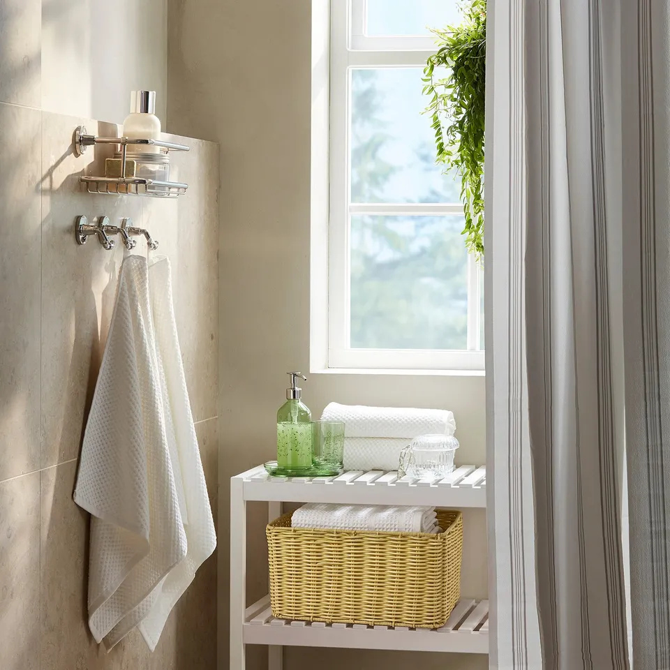 Handwoven yellow basket on shelf, holding towels, under window with plant. White towel on chrome rack, striped shower curtains.
