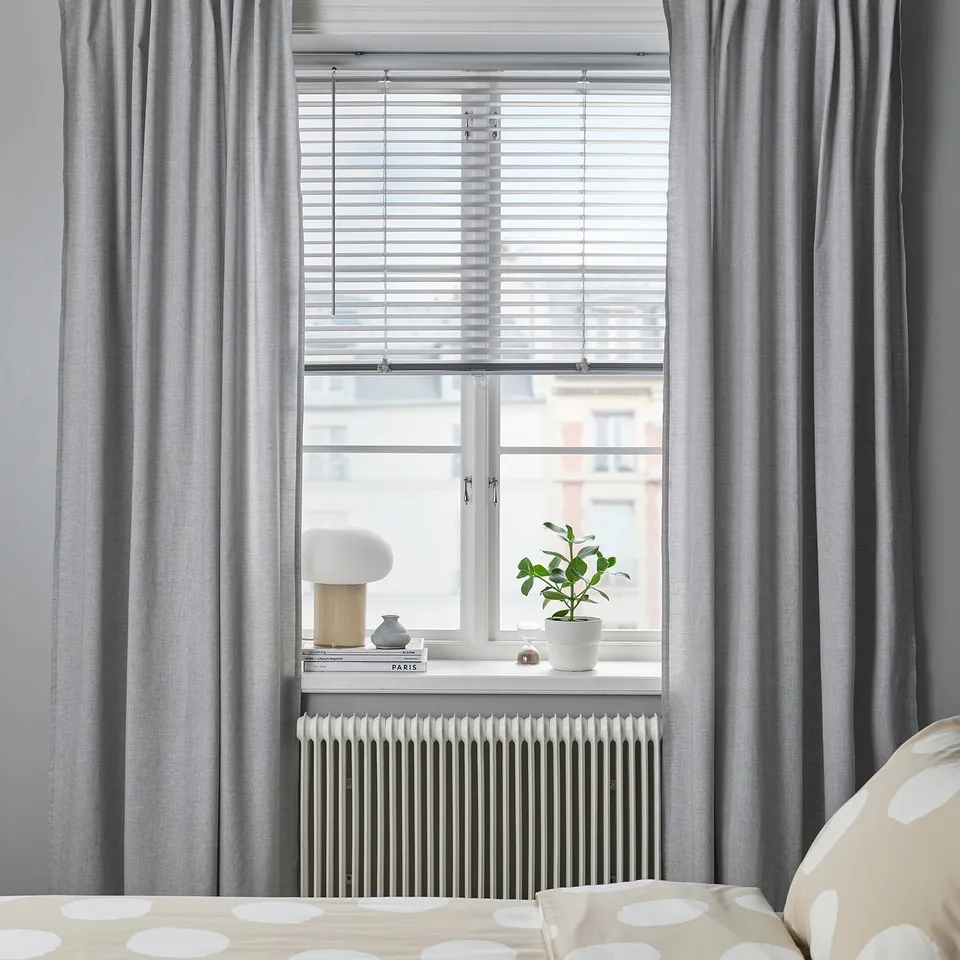 Modern bedroom window with VECKLARFLY blinds, grey curtains, and a white radiator. Decor includes a plant, books, and lamp.
