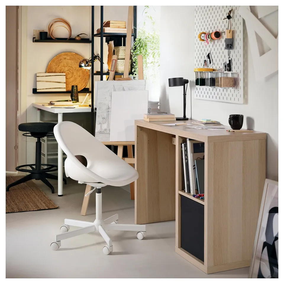 Home office with KALLAX light wood desk, white chair, black lamp, and organised shelves.