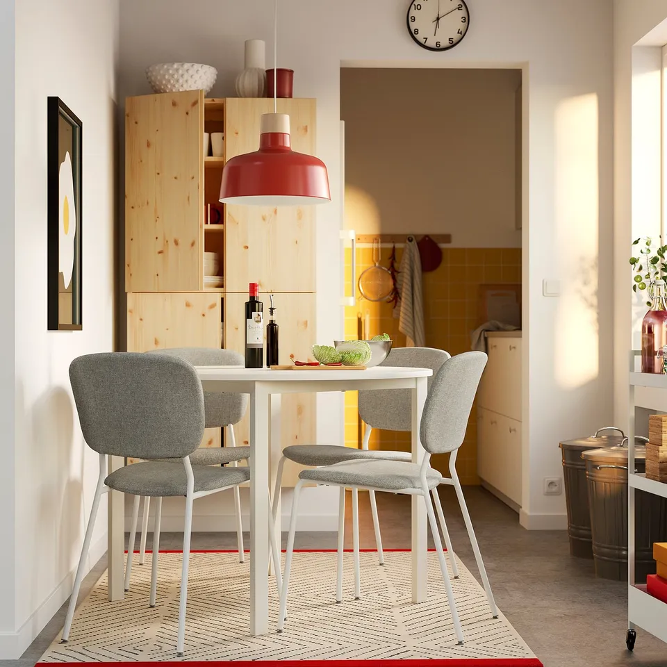 Modern dining area with small white table and grey chairs. Red lamp above, wooden storage behind, kitchen in view.