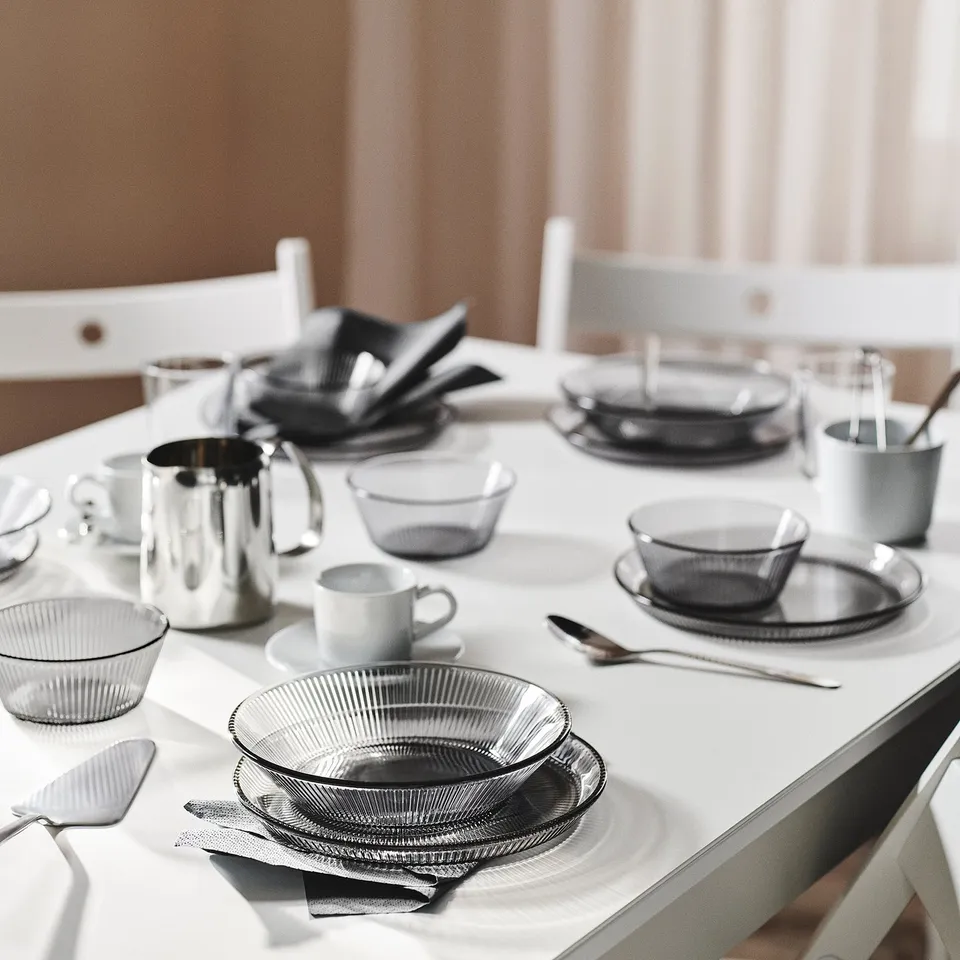 White table set with HAVSGÄDDA grey glass bowls, cups, and cutlery. Bowls are small, round with linear design, stacked for space-saving.
