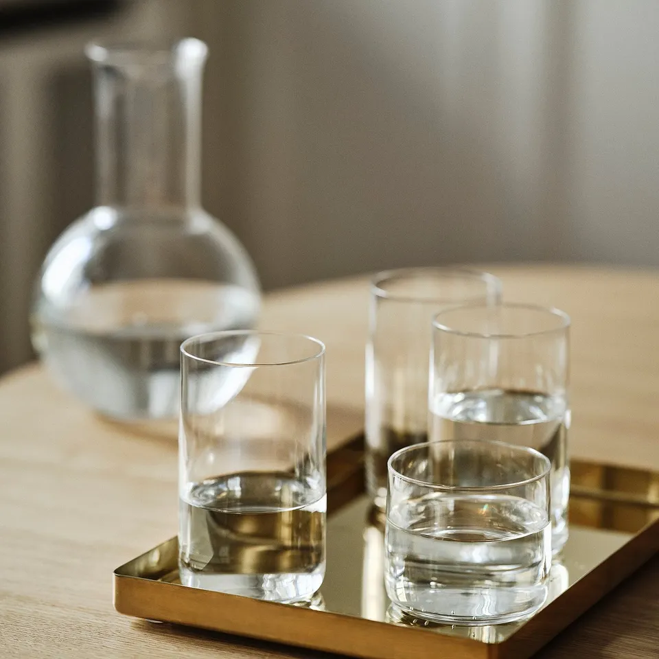 Three clear glass cups, two stacked and one filled with water, on a gold tray beside a pitcher.