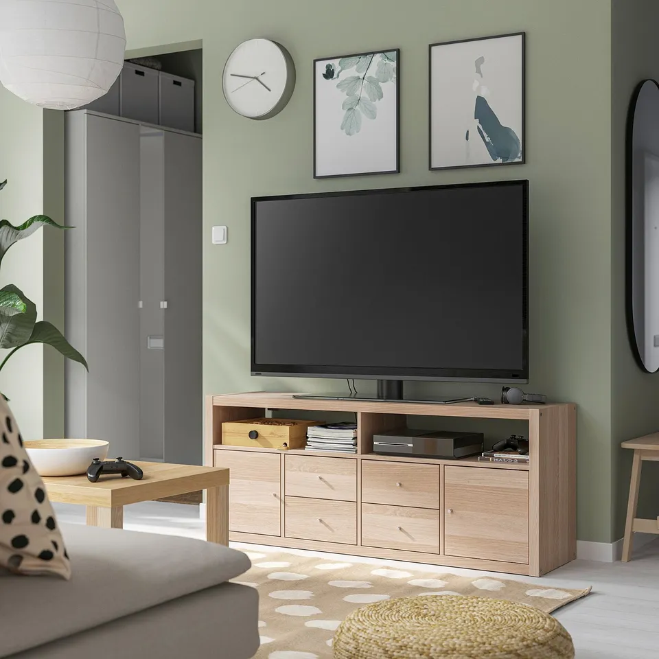 Modern living room with flat-screen tv on KALLAX stand, gaming consoles, art prints, grey sofa, and green walls.