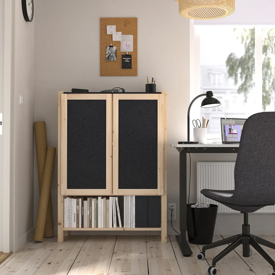 A modern home office an IVAR storage unit with black felt doors, a black office chair, and a wooden desk by a window.