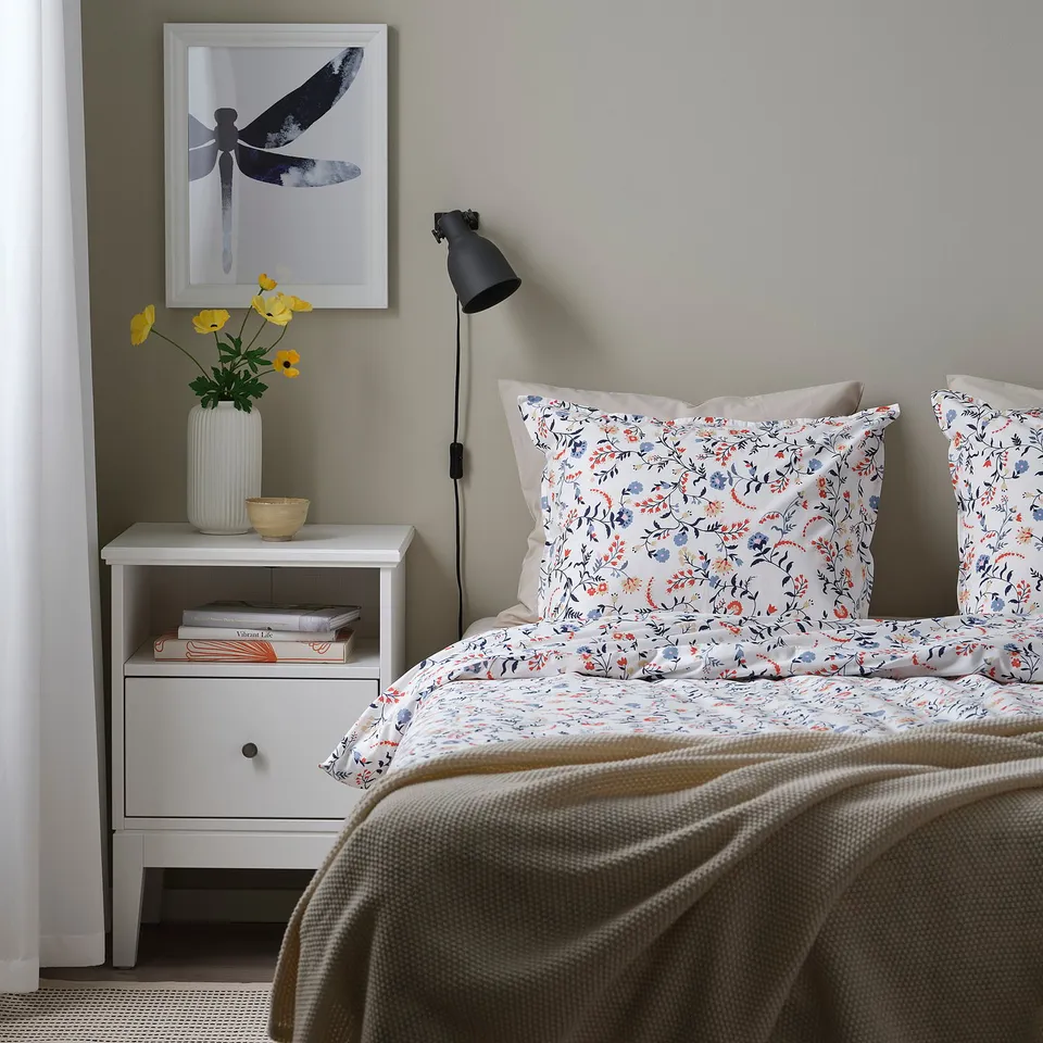 Bedside table with flowers, lamp, and partially visible bed; bed has floral duvet, pillows, blanket in neutral colours.