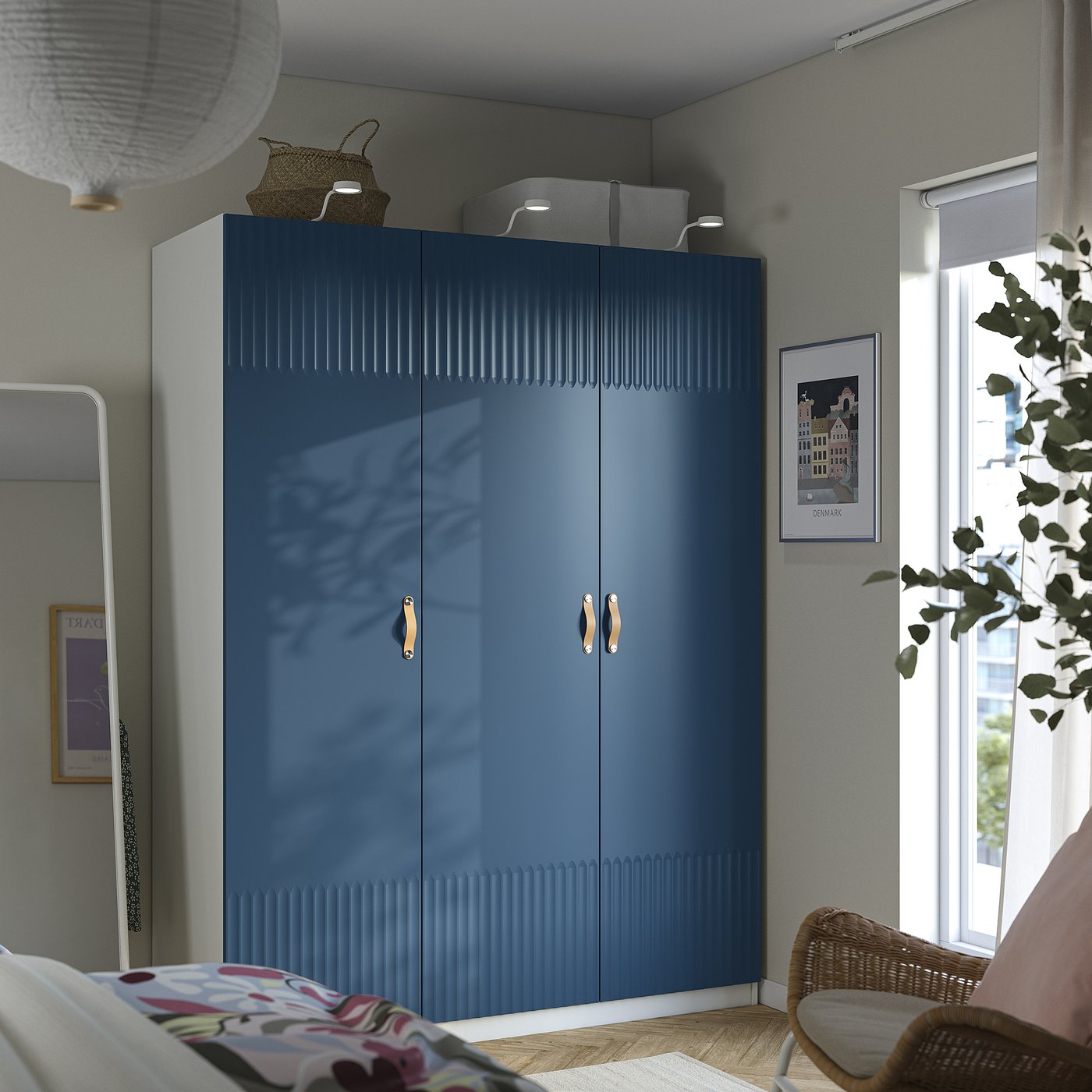 Modern bedroom with blue PAX wardrobe, storage baskets, wicker chair, mirror, floral bed, and hanging plant.