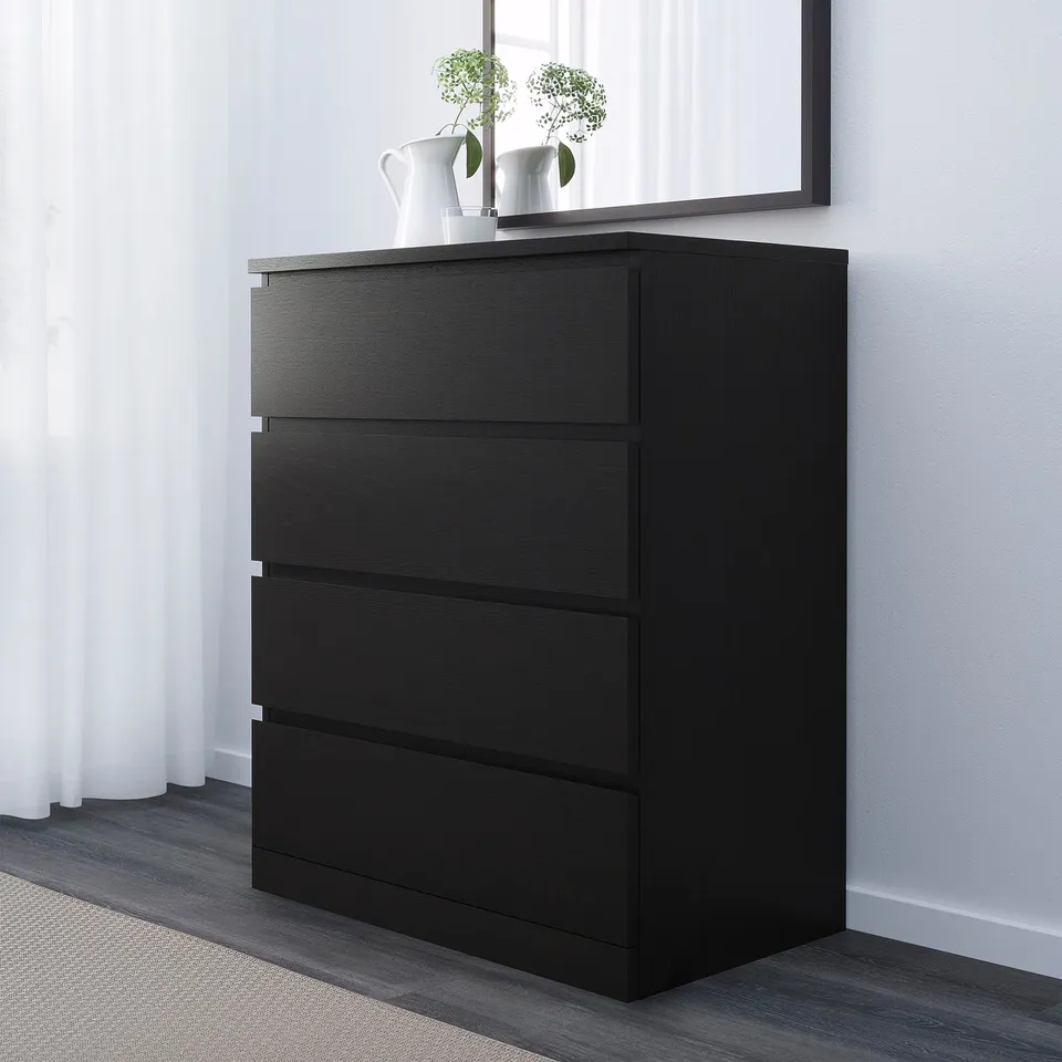 Black chest of 4 drawers against white wall with mirror and flowers on top.