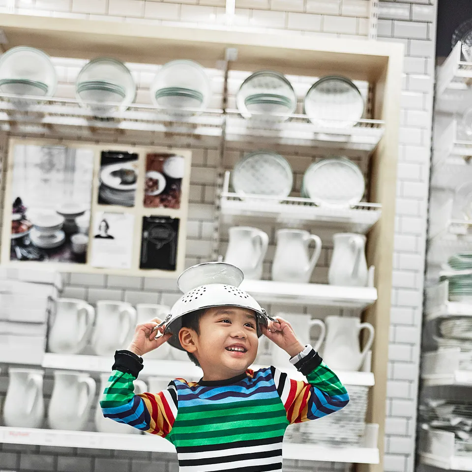 A young child wears a white colander as a hat in an aisle of dishes, smiling happily. The child is wearing a colourful striped shirt.