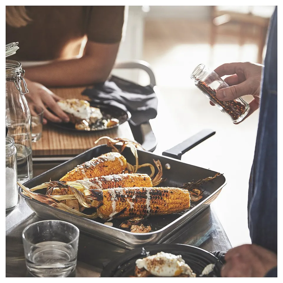 Cooking scene: corn on grill pan, person seasoning, plates, cup, and glasses on table.