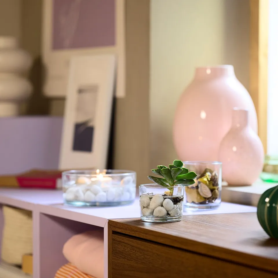 Small glass containers on shelves: one with rocks and a plant, another with white pebbles and a lit candle. A large pink vase is also seen.