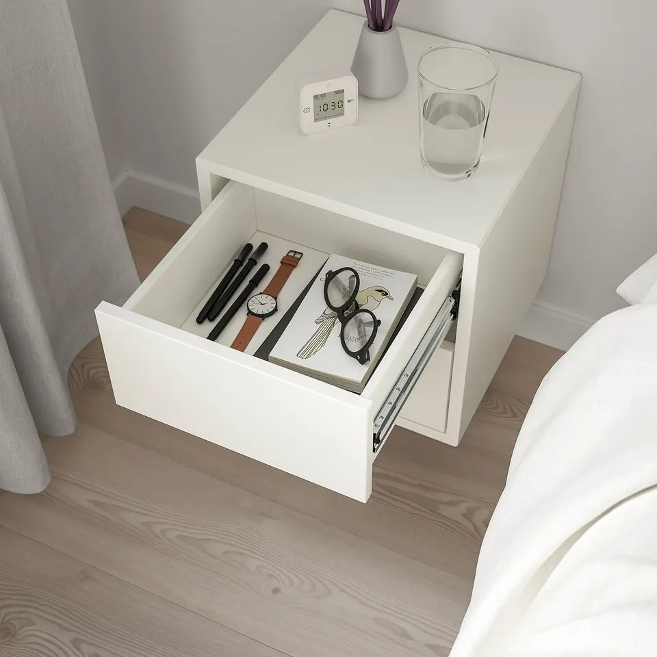 Small white bedside table with open drawer, showing items inside. Clock, glass, and vase on top.
