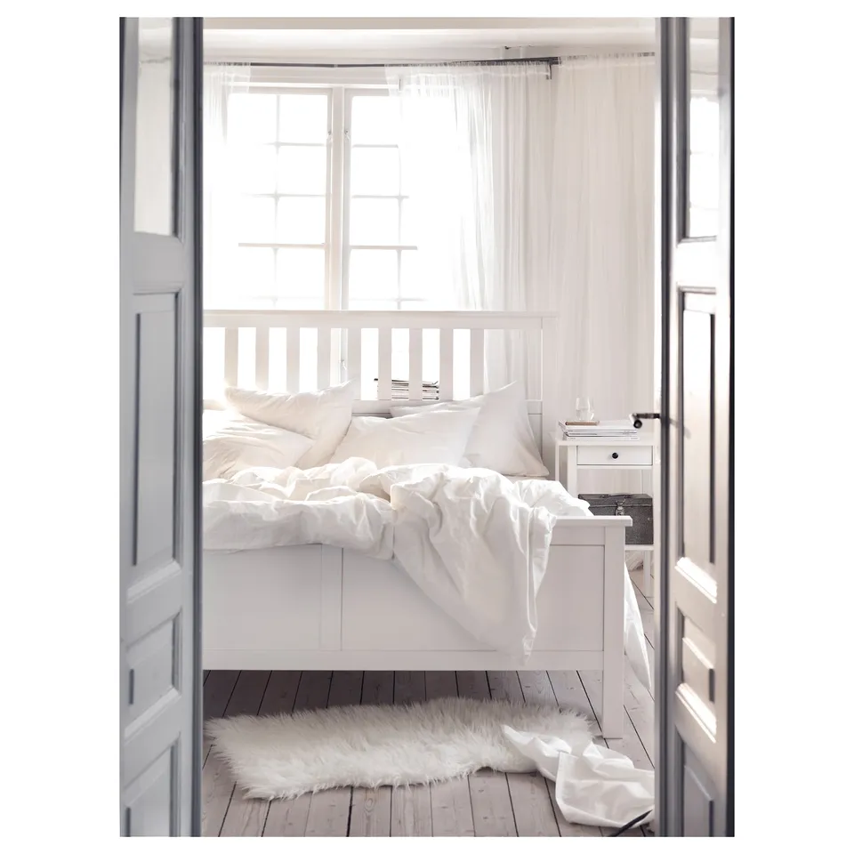 White wooden bed framed with textured wall & large window. Soft white linens, fluffy rug beneath, cosy & bright.