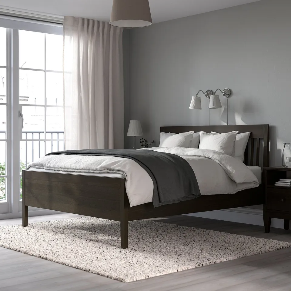 IDANÄS bed frame, dark brown, near window. Classic design, sturdy spindles. Grey walls, white lamp, fluffy rug.
