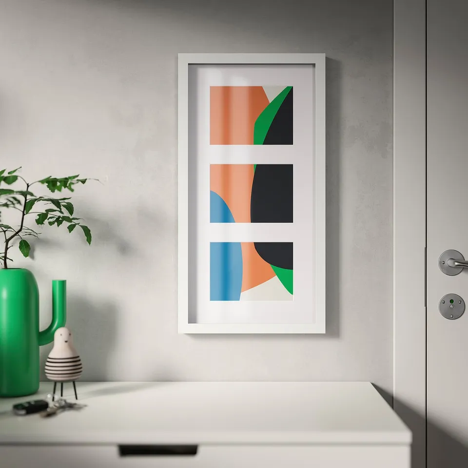 Frame displays three abstract colour blocks; hangs vertically on wall above white drawer, next to green vase with plant.