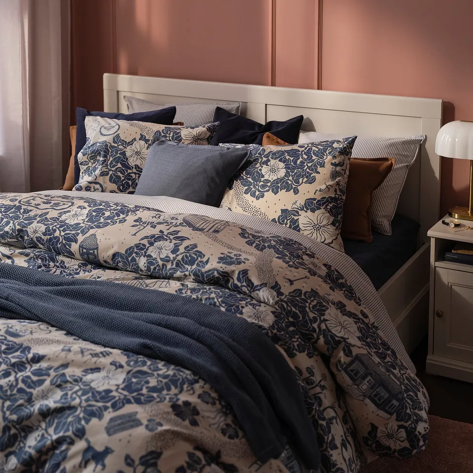PJÄXE blue floral bedding set on large bed, matching the pink wall, with diverse pillows, bedside table, and lamp, creating cosy atmosphere.
