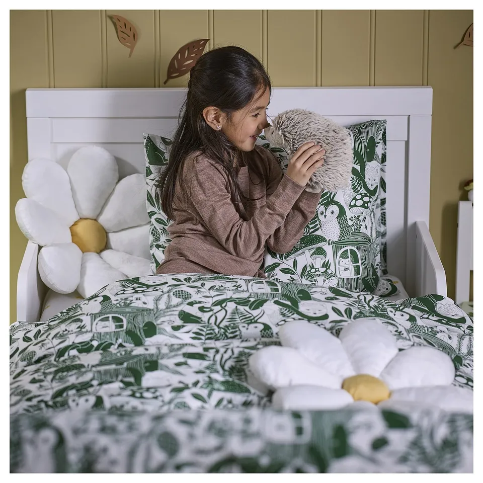 A girl sits on a bed holding a small plush hedgehog. The bed has a green and white forest-themed duvet cover, matching pillows, and a stuffed flower.
