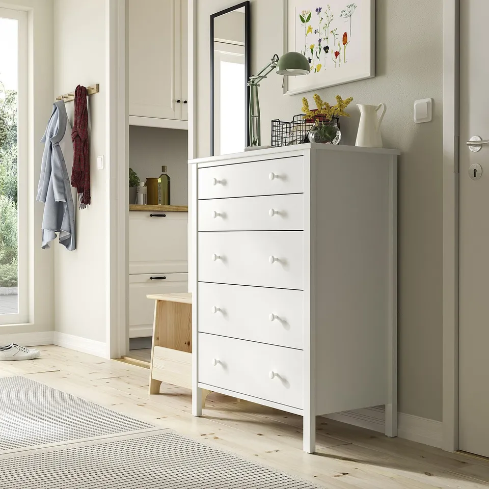 White chest with 5 drawers, tall, simple design. Near hooks, stool, narrow cabinet, rug.