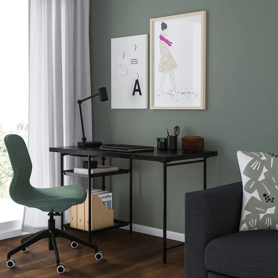 A cosy home office with black desk, turquoise wall, green chair, lamp, and wicker basket. Art above, grey sofa to the right.