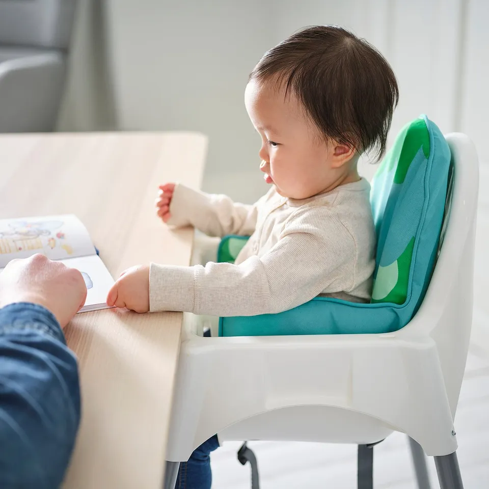 A baby seated in a white highchair with a greenish-blue inflatable cushion for support. The child is reaching for a book held by an adult.