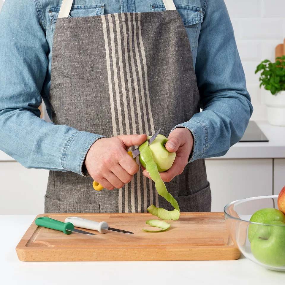 Person peels green apple using UPPFYLLD paring knife on wooden cutting board. Bowl, knife set, and plants nearby.