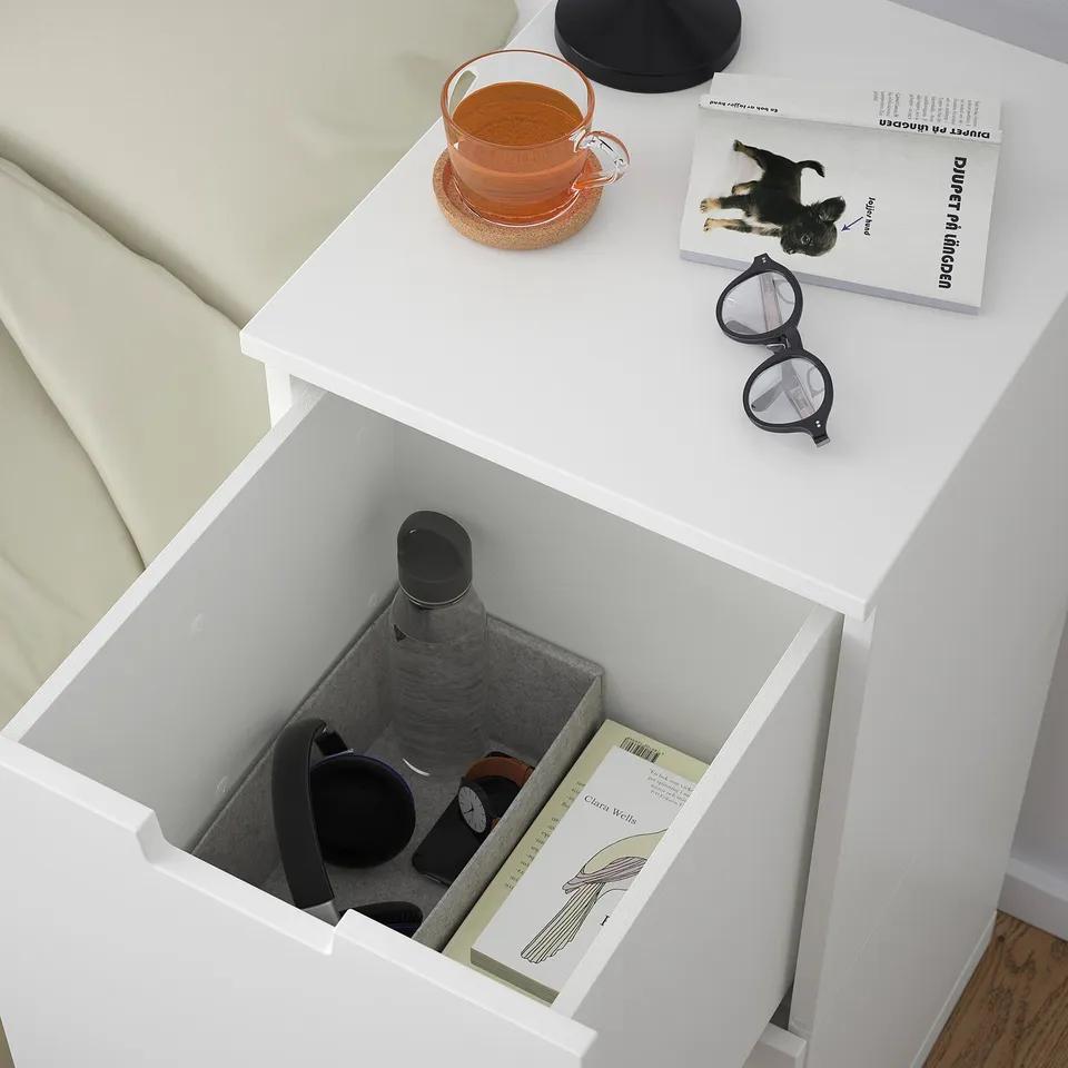 Open bedside table drawer revealing a water bottle, phone, headphones, and book. Top has tea, glasses, and book.