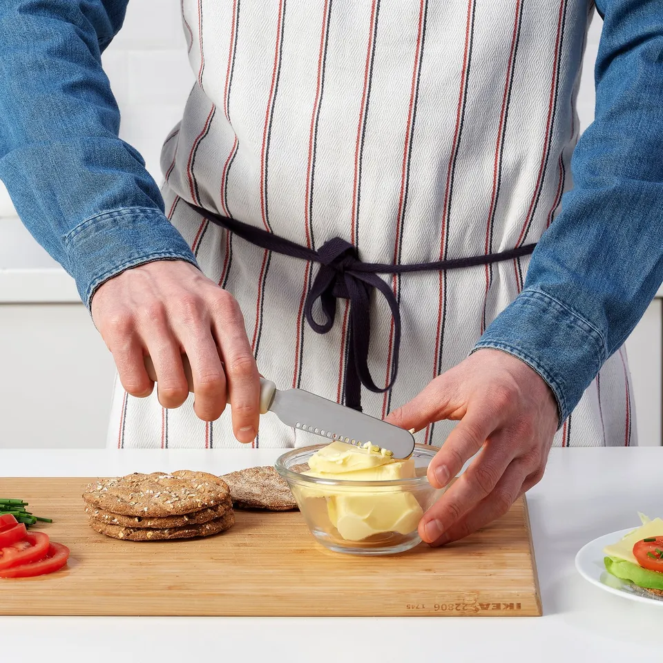 Person spreads butter using a small kitchen tool, with ingredients and sandwich on a wooden board.